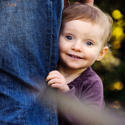 girl peeking around dad's leg and smiling at camera