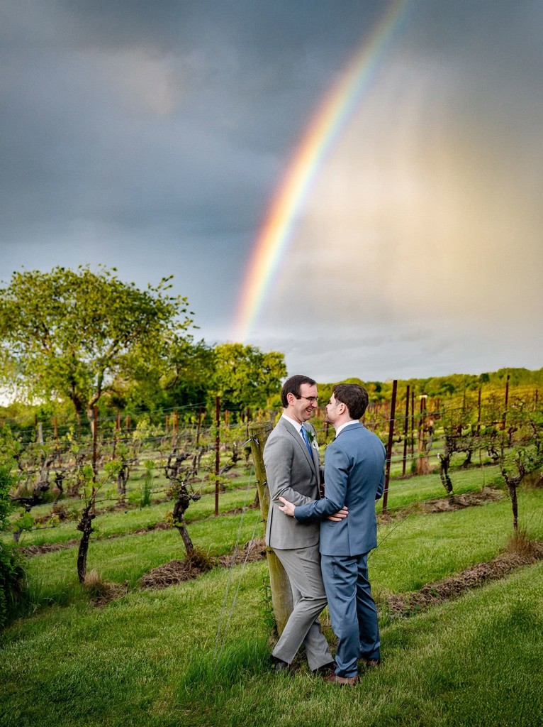 A same-sex couple embraces in a vineyard beneath a rainbow