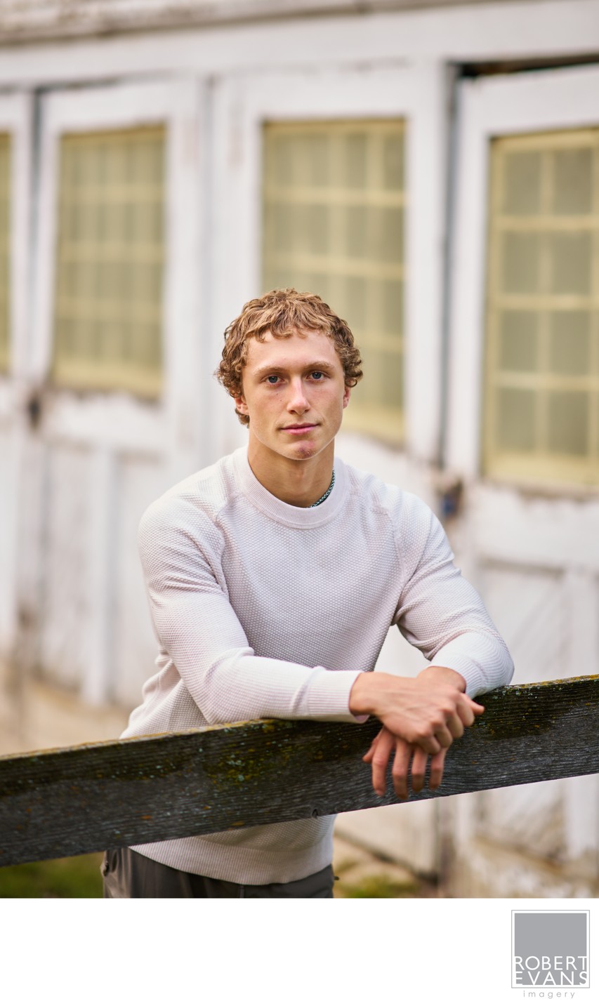 Senior portrait of a boy leaning on a fence post with weathered white wooden doors