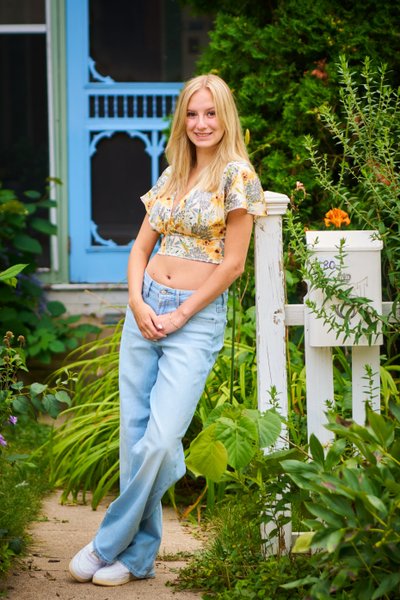 Casual outdoor portrait of a female high school senior leaning against a weathered white picket fence