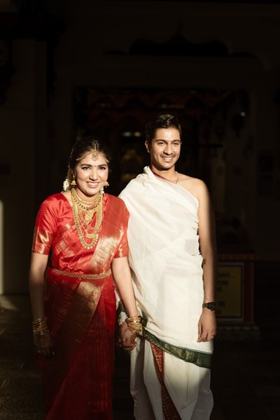 Vidya  in a red silk saree and Sidharth in a traditional veshti holding hands during a South Indian wedding ceremony at Sri Mariamman Temple, Singapore
