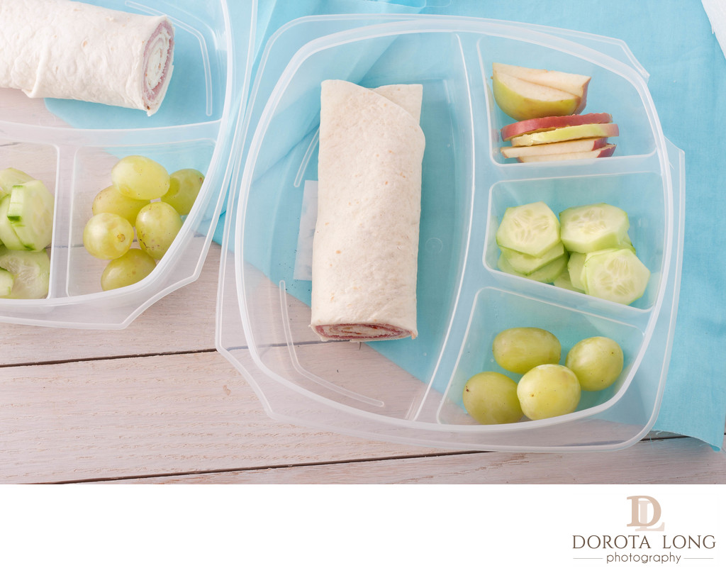 lunch box consisting of a wrap with ham and cheese, apples, grapes and sliced cucumber
