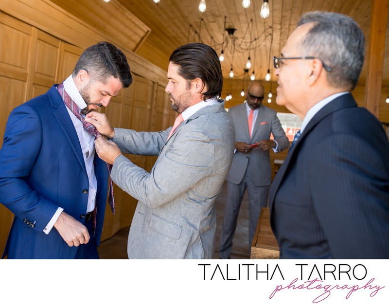 Best man of the groom helps tie a tie while the father of the bride looks on in the barn at Blame Here Ranch. Talitha Tarro Photography