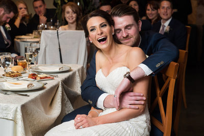 Her new husband embraces her while they sit at their sweetheart table at Knowlton Mansion while listening to at toast during their wedding reception. 