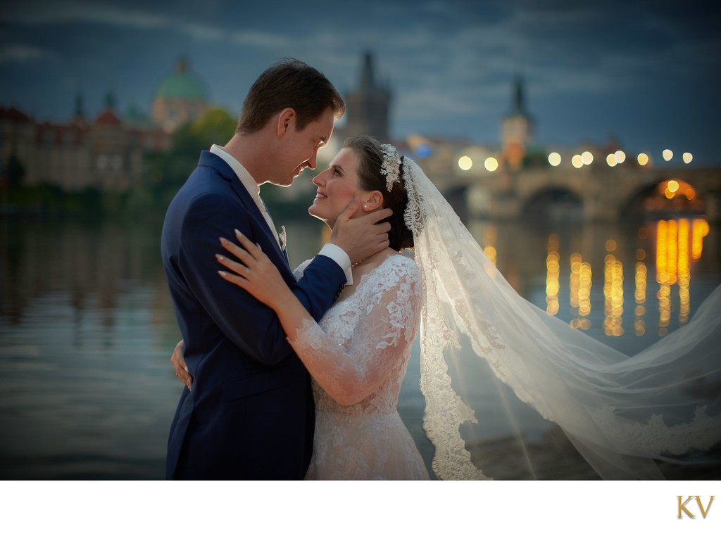 Groom Caressing Bride At Riverside At Night In Prague Near Charles Bridge