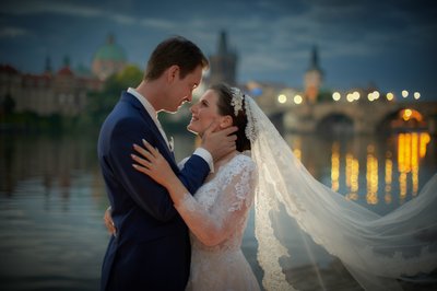 Groom Caressing Bride At Riverside At Night In Prague Near Charles Bridge