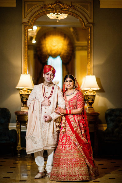 south asian bride and groom standing in front of elaborate mirror smiling